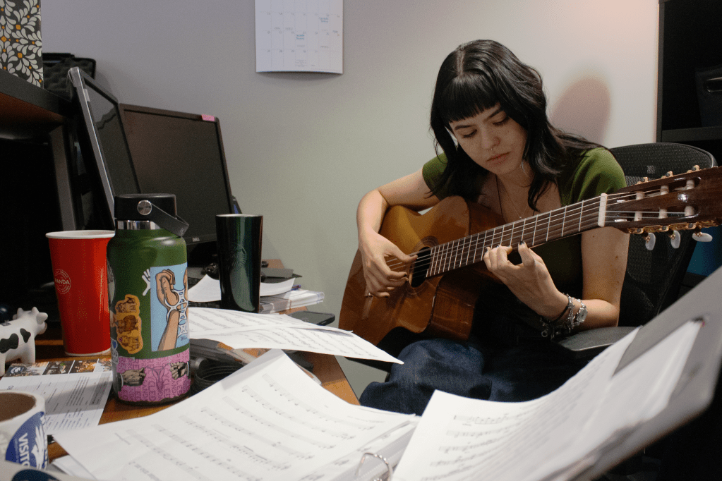 Peyton sits at a computer desk, holding a guitar, surrounded by music sheets and other personal items.